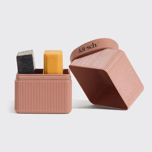 A terracotta, ribbed rectangular container with a matching lid slightly open and leaning against it. The lid features the word “kitsch” in black text. This TSA-approved KITSCH Two Compartment Travel Case is perfect for keeping your beauty essentials organized on the go.