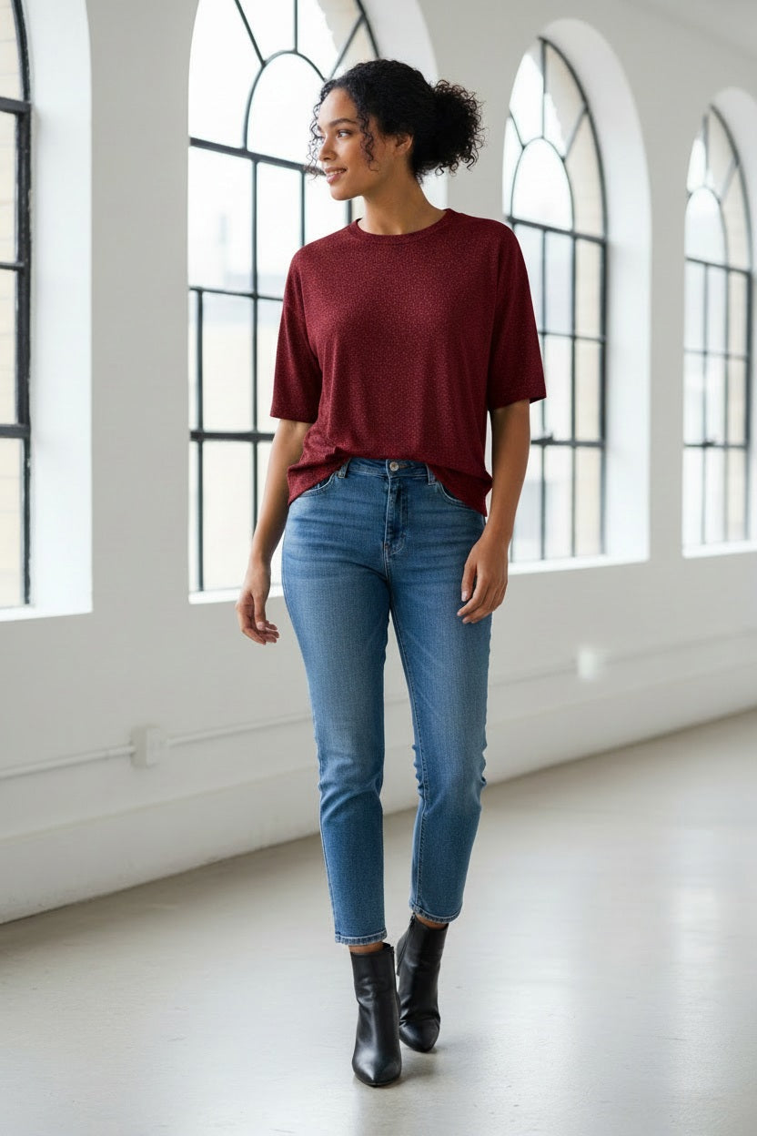 A woman with long brown hair wears the MOMONI - IORA LUREX JERSEY T SHIRT in gold, paired with beige and blue wide-leg floral pants and brown sneakers, standing against a neutral studio background.