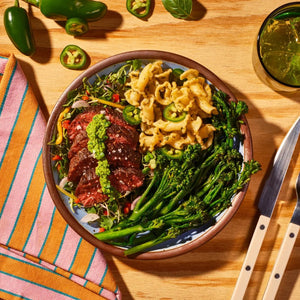 A plate featuring sliced steak generously topped with SAUZ - BASIL JALAPENO PESTO by SAUZ, served alongside broccolini, jalapeño mac and cheese, and microgreens. The wooden table is set with a striped napkin, utensils, fresh jalapeños, and a drink.