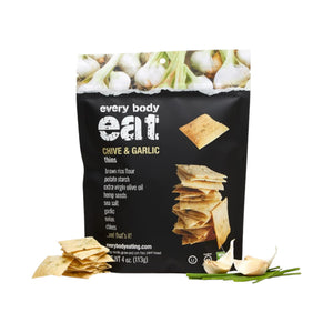 A bag of EVERY BODY EAT CHIVE & GARLIC SNACK THINS from EVERY BODY EAT is pictured alongside gluten-free crackers, garlic cloves, and chives. The black packaging features images of crackers and garlic bulbs at the top, a list of ingredients, and highlights these allergy-friendly snacks.