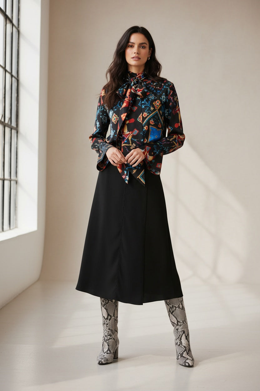 A person with dark hair is standing against a plain background. They are wearing a long-sleeve blouse with a colorful, intricate pattern paired with an elegant SFIZIO - BLACK SKIRT WITH LACE INLAY by SFIZIO. Their outfit, finished with knee-high snakeskin boots made in Italy, looks chic as they look directly at the camera.
