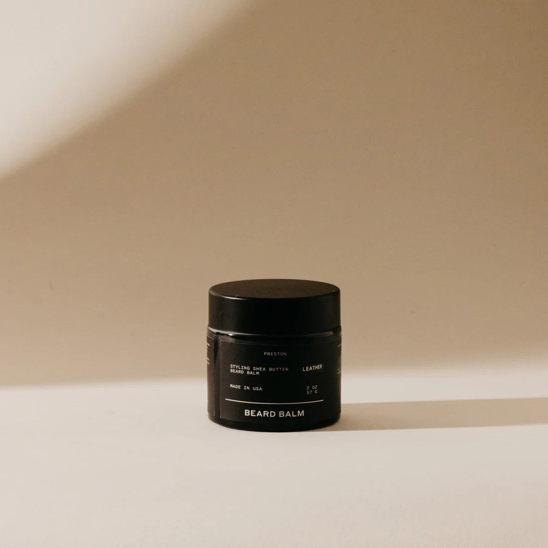 A small black container of PRESTON's cruelty-free beard balm is placed against a neutral, softly-lit backdrop. The label reads "PRESTON - BEARD BALM" along with other details in light/white text. Shadows fall to the right, adding depth to the minimalist design setting that showcases its satin finish.