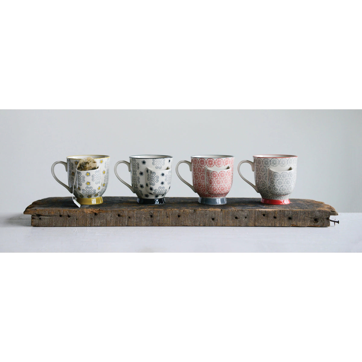 Four intricately patterned stoneware mugs from Creative Coop, each with a unique color, are lined up on a wooden plank against a plain background. Their unique designs create a visually appealing arrangement and, as the &quot;Mug with Tea Bag Holder,&quot; they are microwave safe for practicality.