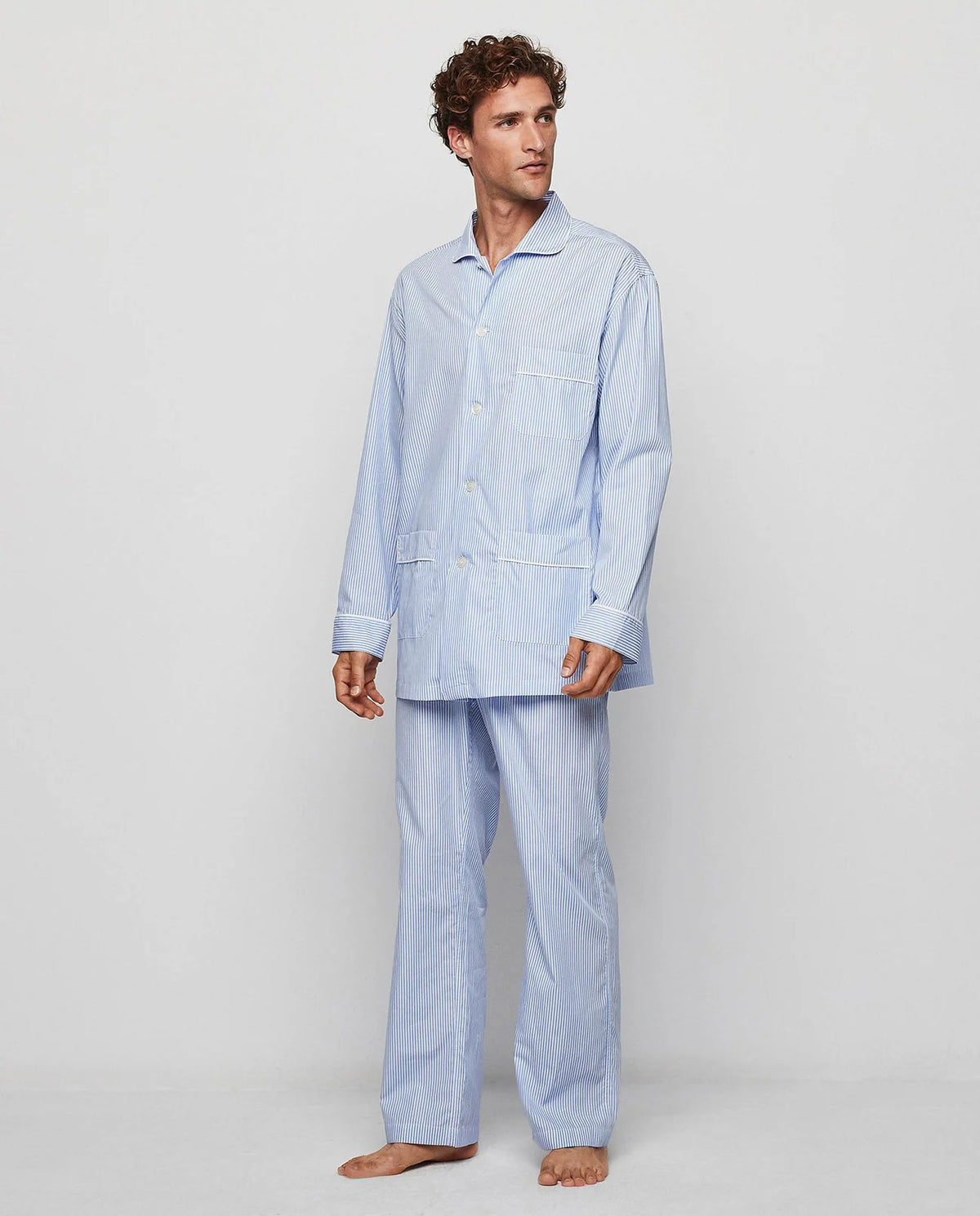 A man stands barefoot facing away from the camera in a MIRTO 1956 - BLUE STRIPE COTTON POPLIN PAJAMA SET, featuring long sleeves and pants—a timeless men’s sleepwear piece by MIRTO 1956, shown against a plain light background.