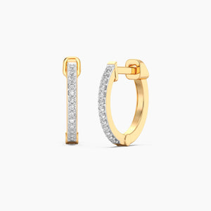 A close-up of a woman’s ear wearing the ELLA STEIN - CLASSIC PAVE DIAMOND HUGGIE EARRINGS (LARGE) in gold, adorned with natural diamonds. Her hand, featuring neatly manicured nails, gently touches her hair near her ear.