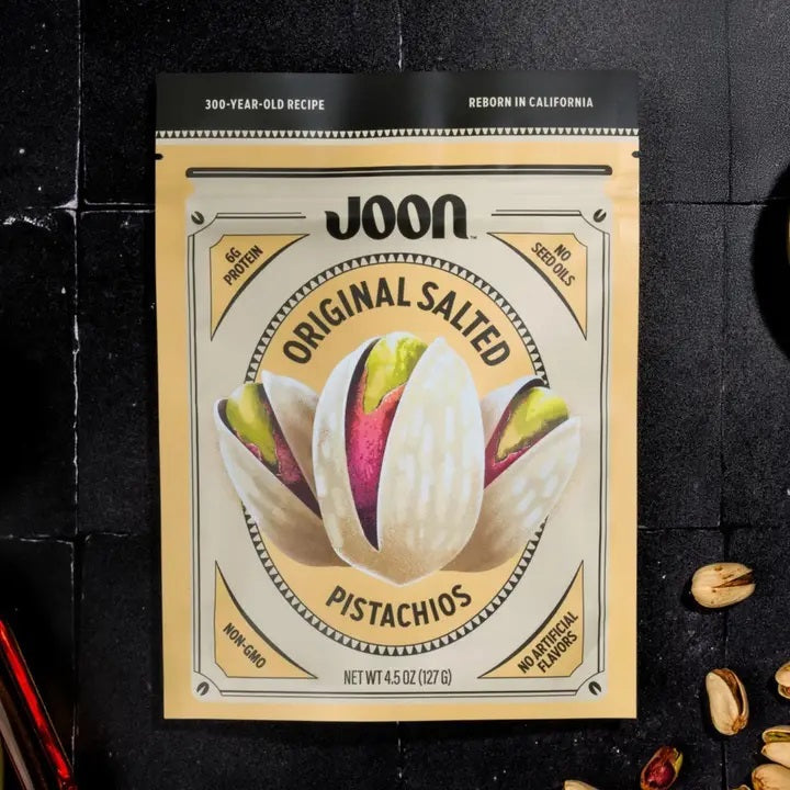 A bag of JOON Original Salted Pistachios, a distinguished offering from the JOON brand, rests on a dark surface with several shelled and unshelled pistachios scattered around. The packaging emphasizes “No GMOs” and “No Artificial Flavors,” underscoring these premium nuts as an irresistible gourmet snack.