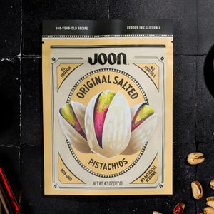 A bag of JOON Original Salted Pistachios, a distinguished offering from the JOON brand, rests on a dark surface with several shelled and unshelled pistachios scattered around. The packaging emphasizes “No GMOs” and “No Artificial Flavors,” underscoring these premium nuts as an irresistible gourmet snack.