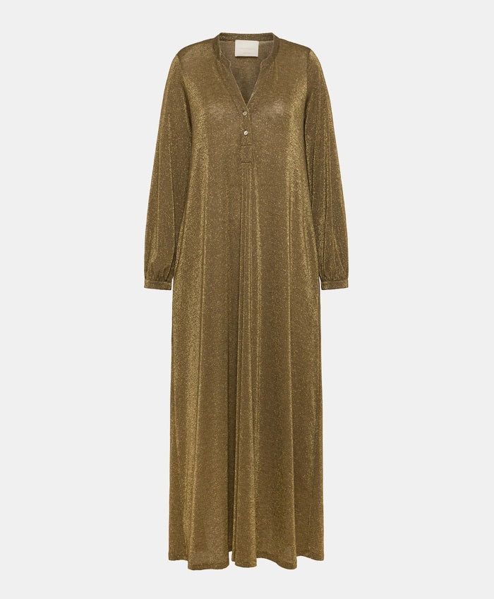 A person stands in a lavishly decorated room, wearing the shimmering long MOMONI - AMEDE DRESS IN LUREX JERSEY, made in Italy by MOMONI, paired with a black wide-brimmed hat adorned with a colorful band. Their hands are in their pockets, and there's a large mirror reflecting part of the ornate wall design.