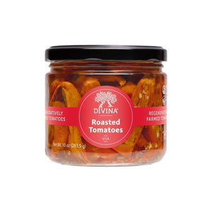 A flatbread topped with lettuce, cucumber, feta, olives, onions, and marinated tomatoes rests on parchment paper next to a jar of DIVINA - ROASTED TOMATOES by DIVINA on a wooden surface.