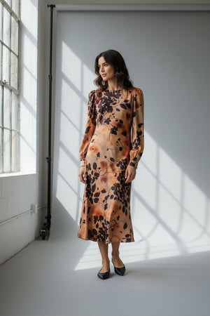 A woman stands with her back to the camera, wearing the MILVA MI - BURNT ORANGE PRINTED DRESS by MILVA—a long-sleeved, viscose midi dress in a rich brown shade accented with an orange and black botanical print—paired with black knee-high boots against a plain light background.
