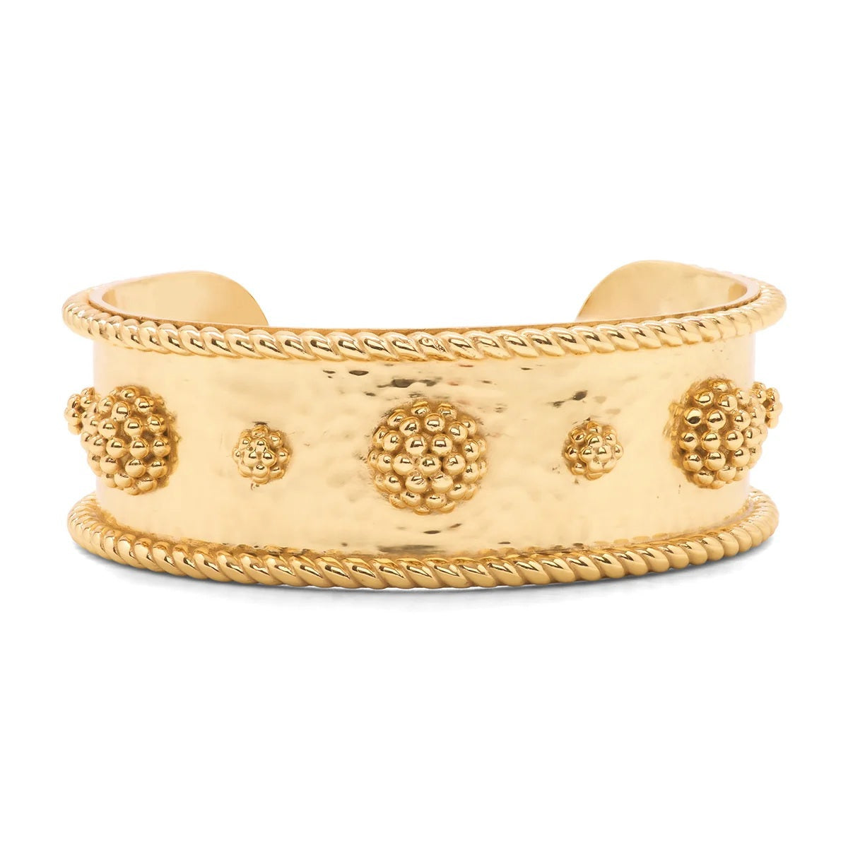 A woman in a white blouse crosses her arms, showcasing several gold bracelets on her wrists, including the bold CAPUCINE DE WULF BERRY CLASSIC CUFF. The softly blurred background features green and yellow tones.