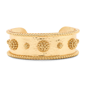 A woman in a white blouse crosses her arms, showcasing several gold bracelets on her wrists, including the bold CAPUCINE DE WULF BERRY CLASSIC CUFF. The softly blurred background features green and yellow tones.