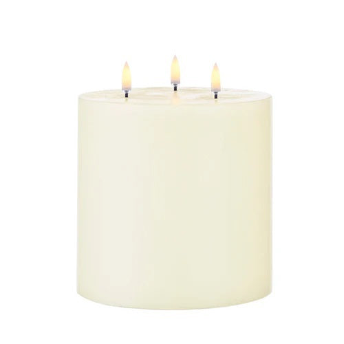 The PIFFANY UYUNI - IVORY TRIPLE FLAME PILLAR CANDLE 6X5.9 features three lit wicks that burn evenly at the top center of the large, ivory-colored candle, set against a plain white background.
