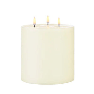 The PIFFANY UYUNI - IVORY TRIPLE FLAME PILLAR CANDLE 6X5.9 features three lit wicks that burn evenly at the top center of the large, ivory-colored candle, set against a plain white background.
