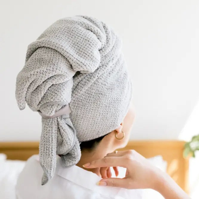 A person with short dark hair, wearing a white shirt, wraps their hair in the VOLO BEAUTY - HERO HAIR TOWEL LUXE and adjusts it with both hands in a bright, airy space filled with soft natural light.