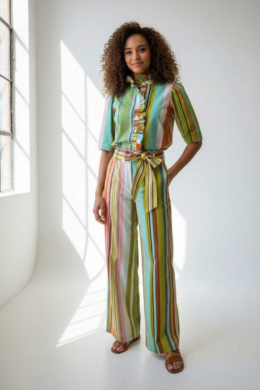 A person models the HOPE FOR FLOWERS Frilled Button Down Shirt in Combo Stripes by HOPE FOR FLOWERS, featuring vibrant vertical stripes and a ruffled collar, crafted from GOTS certified organic cotton for sustainability.