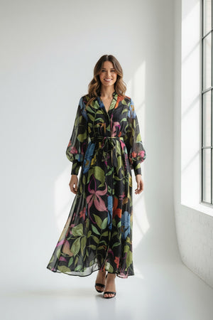 Wearing the LAVI COUTURE - ARTEMISIA FLORAL DRESS, a woman showcases a flowing black dress with vibrant botanical designs in pink, blue, green, and yellow. Against a white background, she highlights its cinched waist and long sleeves while pairing it with black strapped heels.