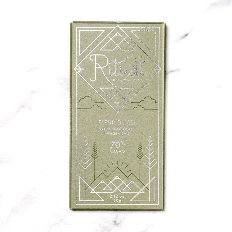 A green RITUAL CHOCOLATE - FLEUR DE SEL CHOCOLATE BAR 70% CACAO with silver geometric designs and text on the wrapper, shown on a white marble background.