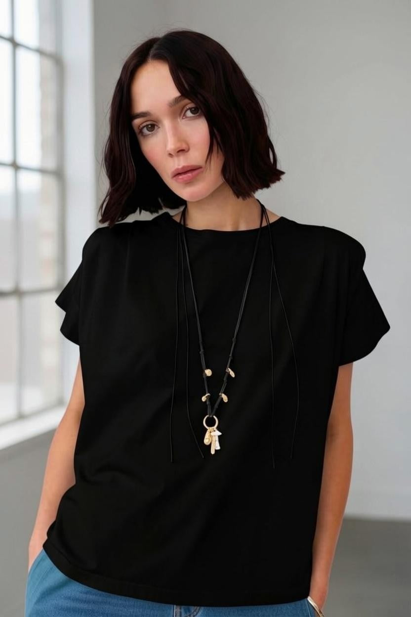 A woman with chin-length dark hair wears the FLOOR - BLACK COTTON TOP by FLOOR, a relaxed and effortlessly stylish piece. She pairs it with blue jeans and a long necklace featuring key-shaped pendants, standing indoors near large windows and gazing at the camera with a neutral expression.