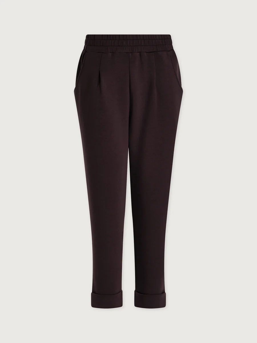A person seen from behind wearing VARLEY - ROLLED CUFF PANT 25” IN COFFEE BEAN dark brown trousers with rolled cuffs and cream-colored ballet flats.