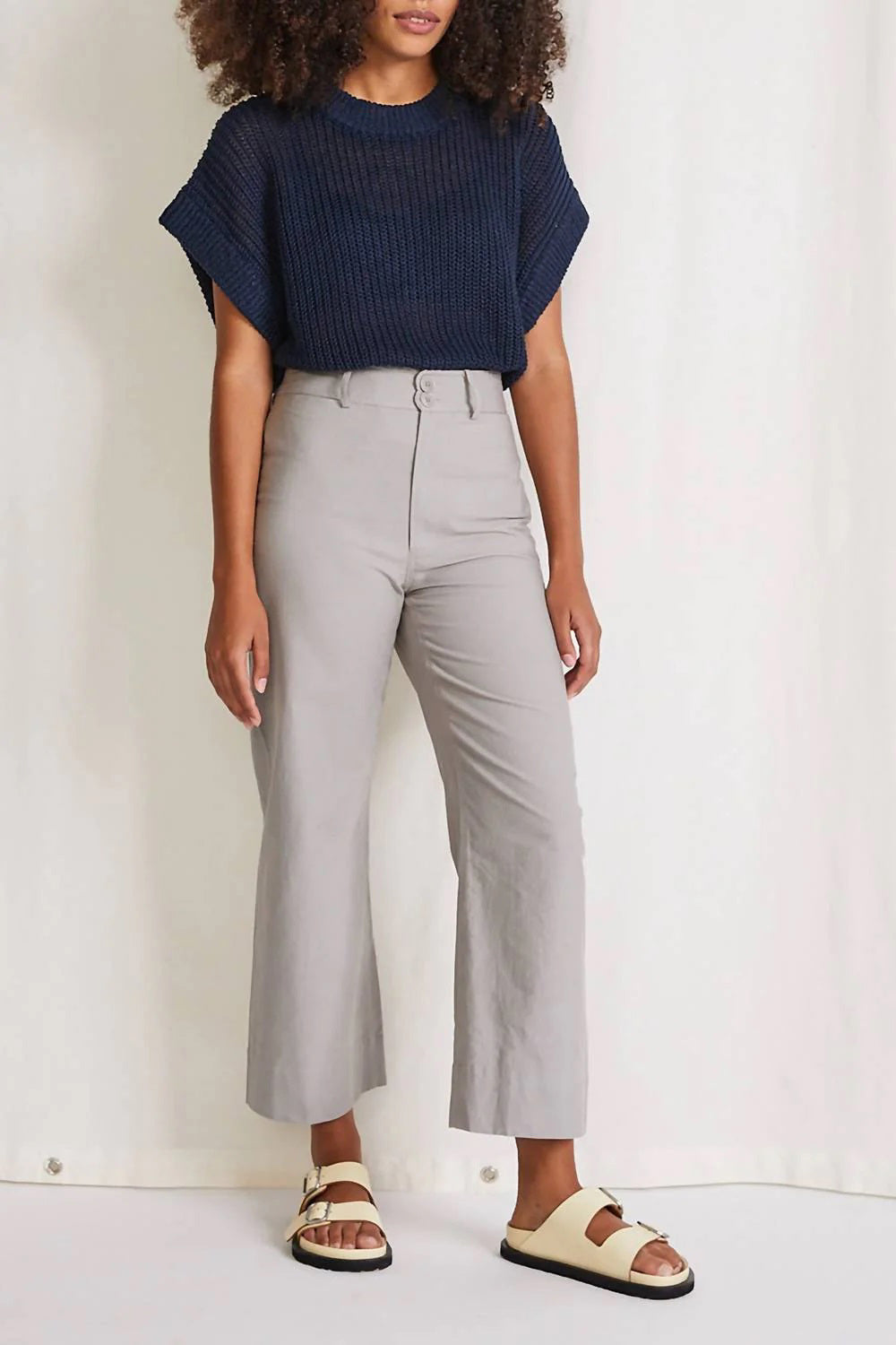 A person is styled in a navy blue short-sleeve knit top paired with the high-waisted, wide-legged CLASSIC MERIDA PANT in light gray by APIECE APART. They stand against a pale backdrop, completing their sleek silhouette with beige sandals. The individual&#39;s face is not visible in the image.