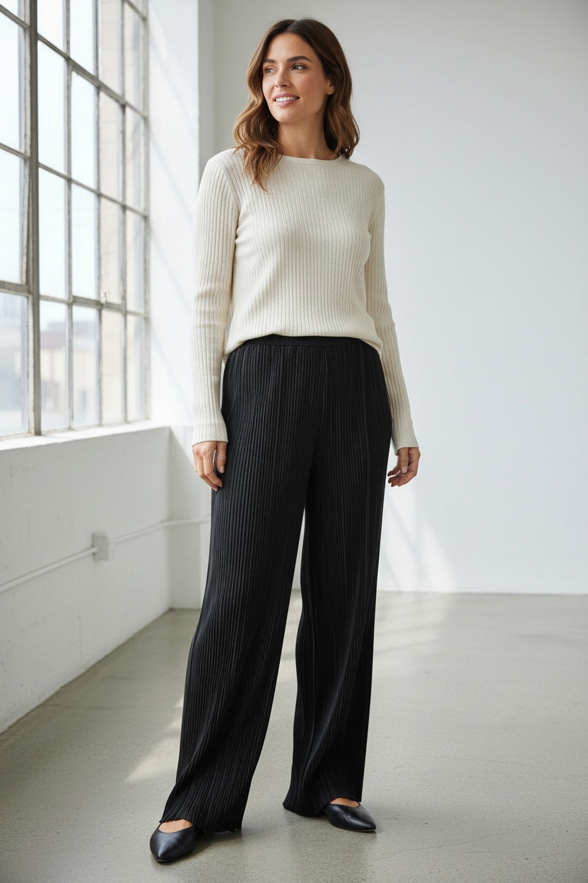 Side view of a person wearing the MEIMEIJ Long Pleated Pant in black, highlighting the elegant texture of pleated fabric. The pants are paired with a light gray, long-sleeved top, and the ensemble's draping is well highlighted against a plain background and light-colored floor.