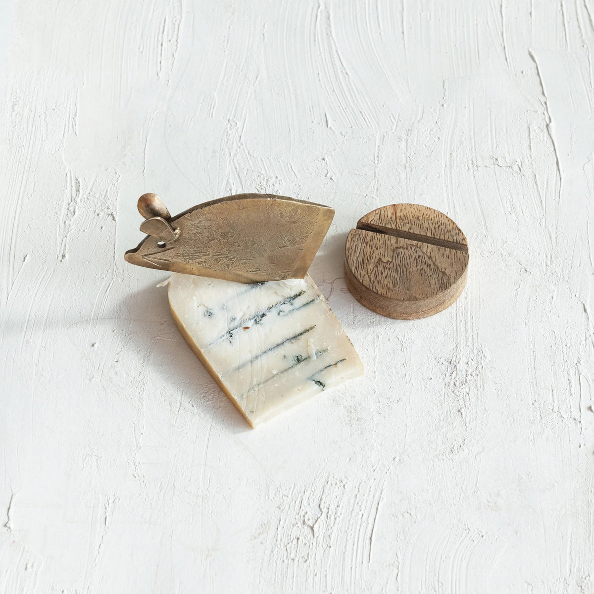 On a textured white surface, a block of blue cheese is accompanied by the Creative Coop Mouse Cheese Knife with Stand and a small, round wooden case with a slot lid.