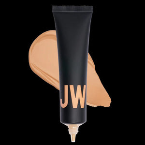 A close-up of a person with natural curly hair, adorned in gold hoop earrings and a white top. They wear a neutral facial expression while looking directly at the camera. The softly lit, plain background enhances their glowing skin as if touched by JASON WU - TINTED MOISTURIZER MEETS CC CREAM from JASON WU BEAUTY.