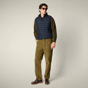 A man wearing the SAVE THE DUCK - RHUS VEST with Recycled Plumtech padding over an olive cardigan, paired with loose-fitting olive pants and brown shoes, stands against a plain light background with his hand in his pocket.