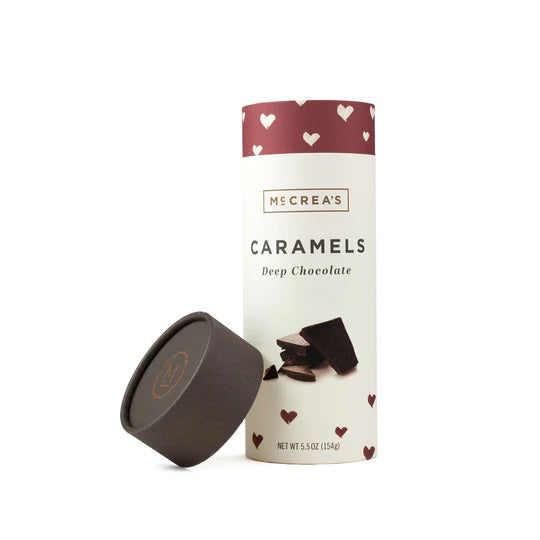A product shot of McCREA'S CANDIES - DEEP CHOCOLATE CARAMELS. The cylindrical container has a maroon lid with a heart pattern and a matching maroon band at the top. The main body is cream-colored with text and images of velvety cocoa pieces. The lid is off and placed to the side, tempting every choco-lover.