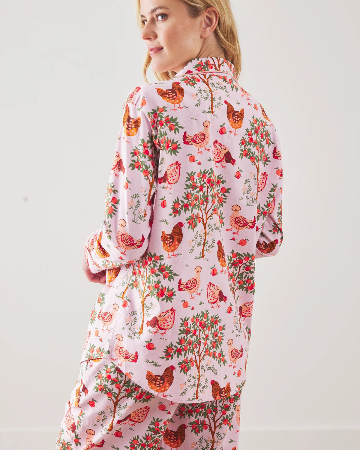 A person stands barefoot against a white wall in PRINTFRESH&#39;s pink organic cotton HEN HOUSE PAJAMA SET, featuring hand screen-printed roosters and floral patterns. The pajamas have long sleeves and pants, with one hand in a pocket and their head slightly tilted.