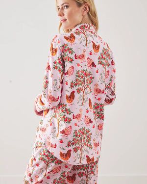 A person stands barefoot against a white wall in PRINTFRESH's pink organic cotton HEN HOUSE PAJAMA SET, featuring hand screen-printed roosters and floral patterns. The pajamas have long sleeves and pants, with one hand in a pocket and their head slightly tilted.