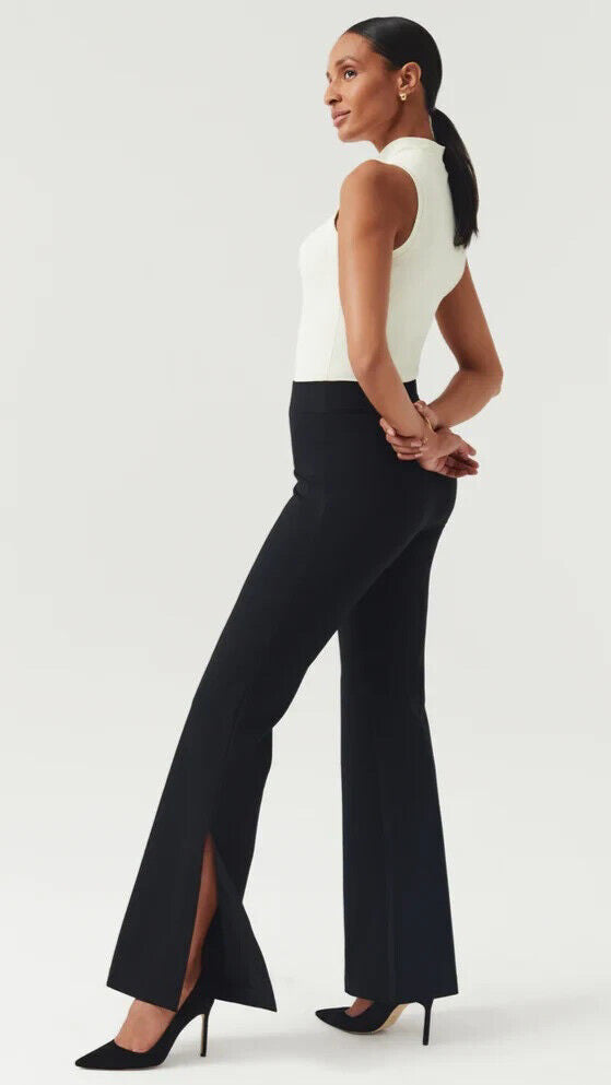 A woman stands in profile against a plain background, wearing a sleeveless white top, black flared pants with front slits, and black high heels. Her hands are clasped behind her back, and her hair is tied back.