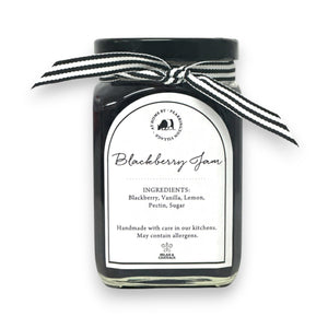 The FEARRINGTON SIGNATURE COLLECTION - FEARRINGTON HOUSEMADE BLACKBERRY JAM features a stylish black and white striped ribbon tied around the lid. The label reveals ingredients including blackberry, vanilla, lemon, pectin, and sugar. This handcrafted jam is made with meticulous care and is an ideal hostess gift. Please be aware that it may contain allergens.