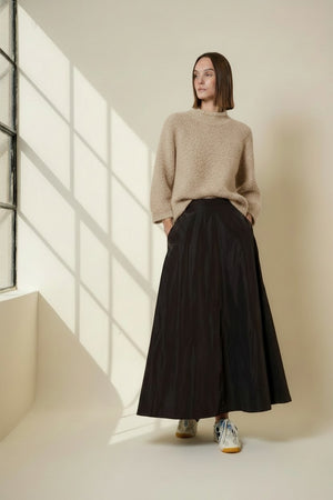 A woman stands indoors by a window, wearing the VICARIO CINQUE - MADEIRA Boucle Sweater in Light Taupe from VICARIO CINQUE, paired with a long black skirt and sneakers. Sunlight casts grid-like shadows on the wall.