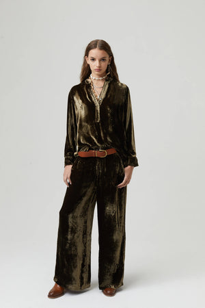 A person with long wavy hair stands against a plain background wearing a mustard yellow, pure silk velvet long-sleeve top and matching, high-waisted LANHTROPY - THE LUNA VELVET PANT by LANHTROPY. They are accessorized with layered necklaces, a brown belt, and flat sandals.