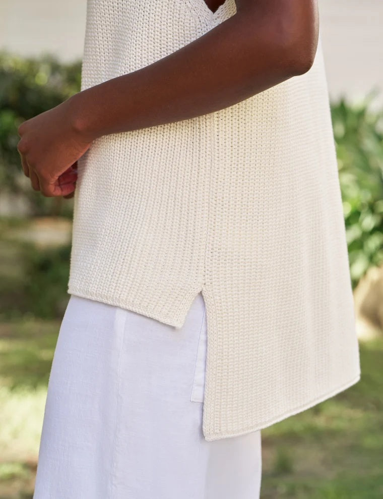 Wearing the FRANK & EILEEN MONTECITO Shell Mock Neck Sweater Shell in Ivory, a person with curly hair and white pants stands outdoors with their back to the camera, in front of a white building surrounded by greenery.