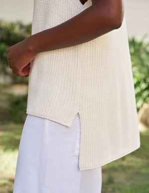 Wearing the FRANK & EILEEN MONTECITO Shell Mock Neck Sweater Shell in Ivory, a person with curly hair and white pants stands outdoors with their back to the camera, in front of a white building surrounded by greenery.
