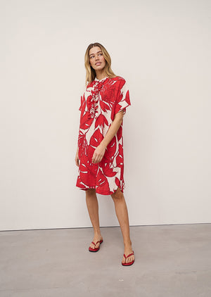 Close-up of the DAISY DRESS IN RED PRINT by 0039 ITALY, featuring a bold red leaf pattern on white viscose blend and vertical ruffles along the button placket. Only part of the woman's face and hair are visible.