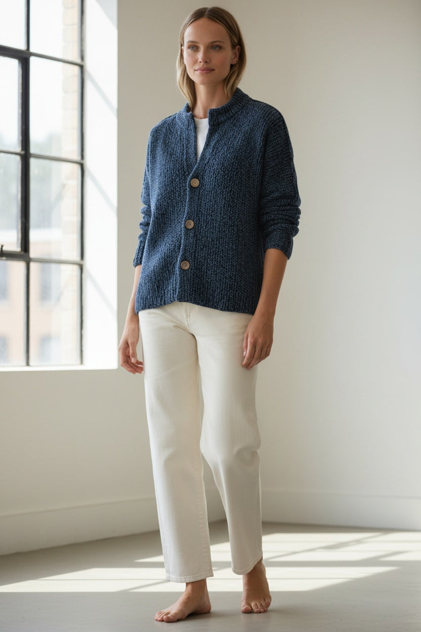 A person with wavy blonde hair, wearing a relaxed fit FRANK &amp; EILEEN Montecito Crew Neck Cardigan in Pure Italian Cotton Blueberry and white pants, stands outdoors facing away, surrounded by greenery and trees.