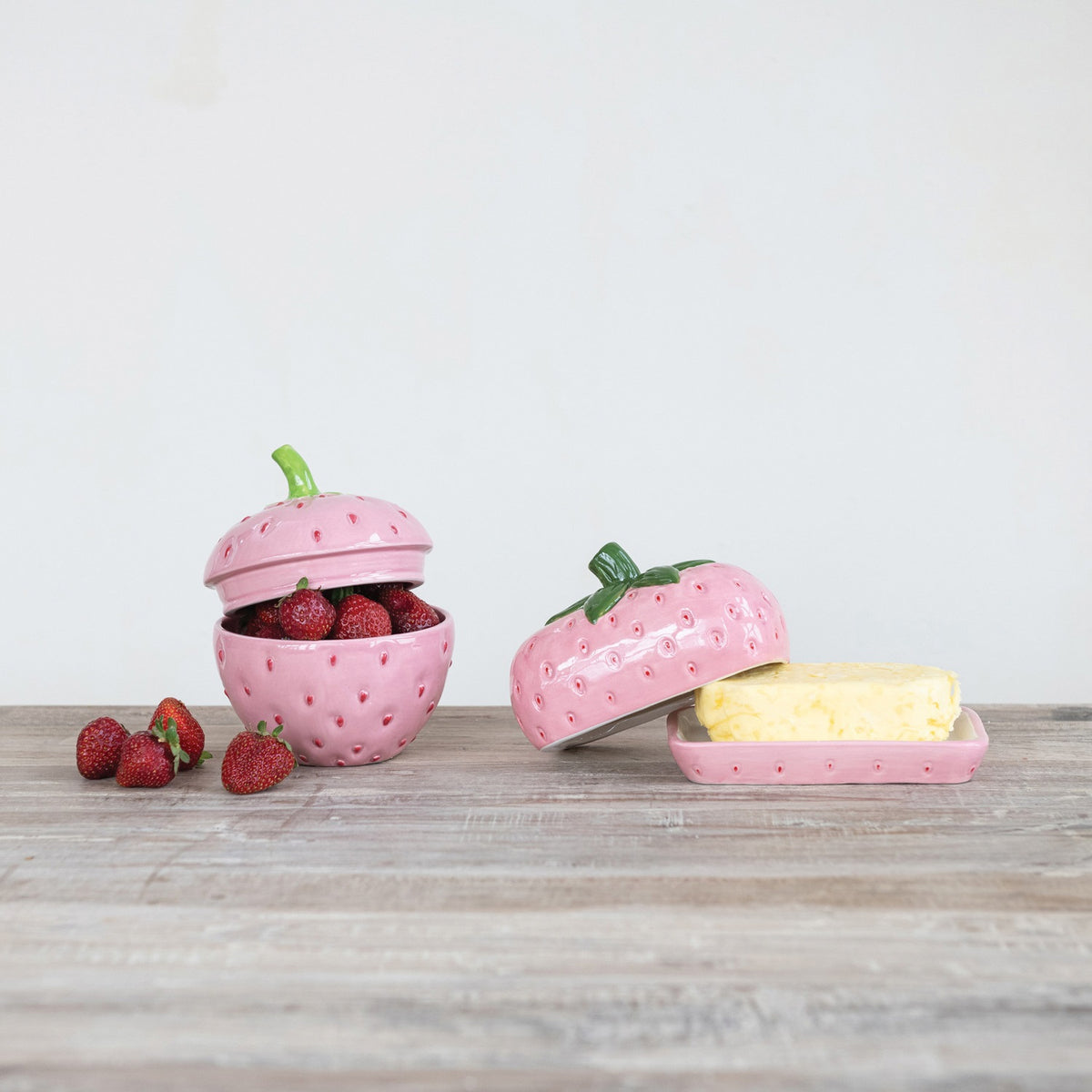 The HAND-PAINTED CERAMIC STRAWBERRY SHAPED BUTTER DISH by CREATIVE COOP features a pink strawberry design with a green leafy top, hand-painted details, and comes on a matching pink rectangular base.