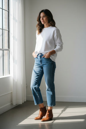A woman with long brown hair wears the FRANK & EILEEN Quinn Long Sleeve Crewneck Tee in Heritage Jersey White, paired with blue jeans, standing in a sunlit kitchen with white tiles and a green stove, smiling softly.