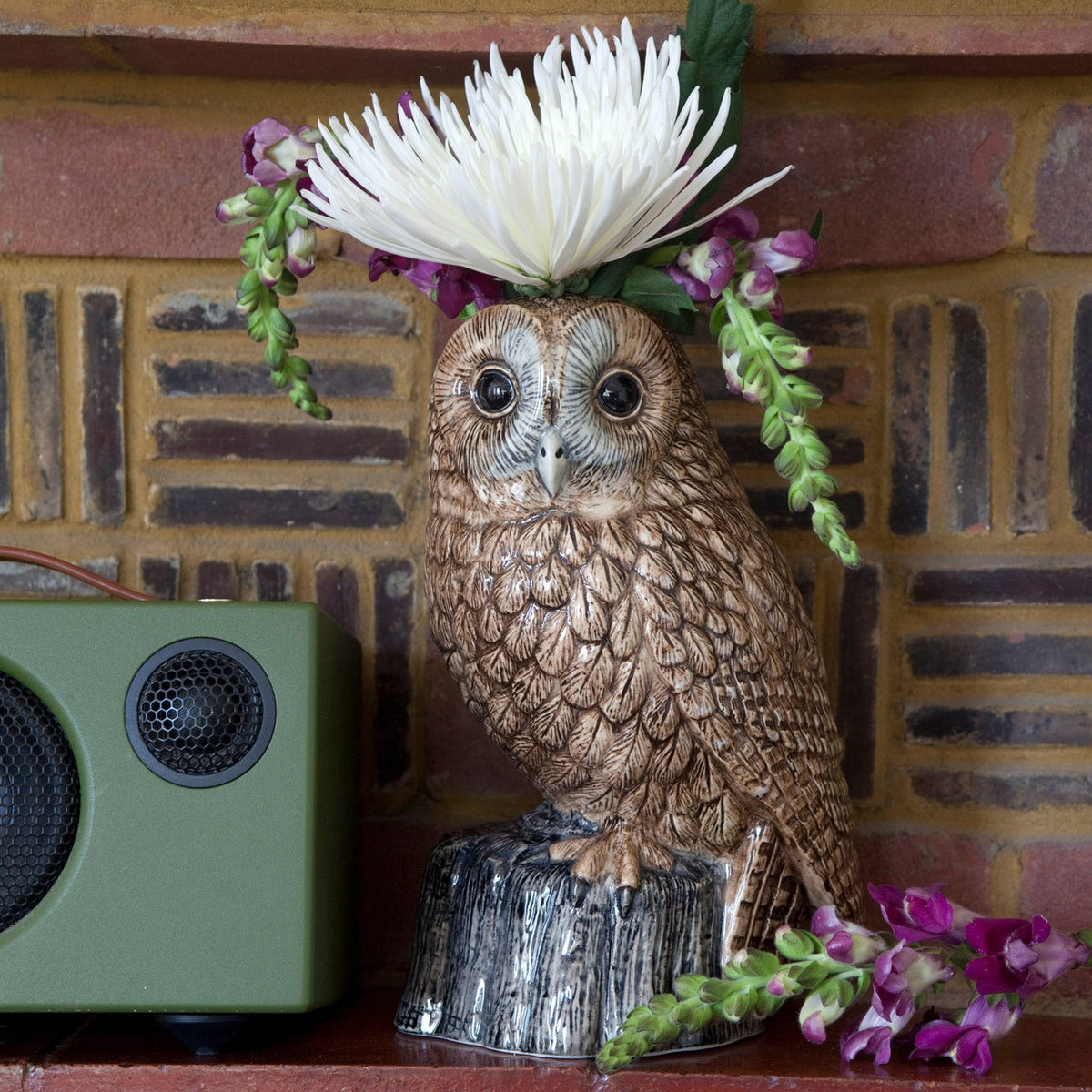 The QUAIL CERAMICS - TAWNY OWL FLOWER VASE by QUAIL DESIGNS features a hand-painted stoneware owl atop a stump-shaped base, with green leaves and white flowers sprouting from its head, set against a plain white background.