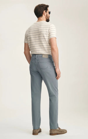 A man stands with his back to the camera against a plain light background, wearing a beige striped short-sleeve shirt, 34 HERITAGE Charisma Relaxed Straight Leg Pant in Flint Stone, brown suede shoes, and sunglasses.