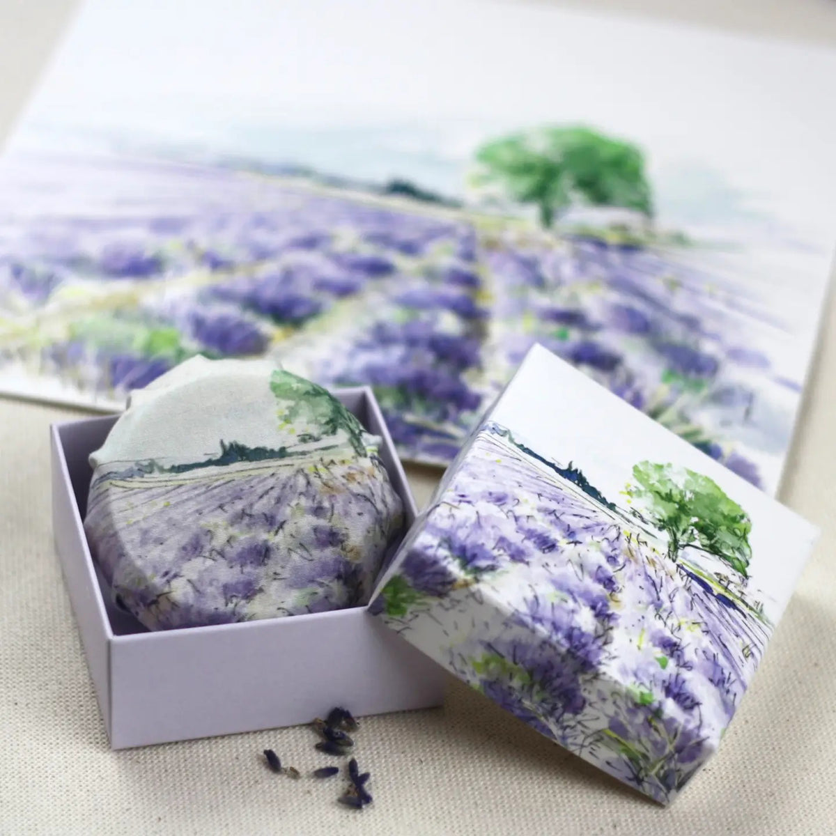 L&#39;AUGUSTE PROVENCE – THE ESSENCE OF PROVENCE Lavender Soap, a round handmade French soap wrapped in cloth with a lavender field and tree motif, rests in an open L&#39;AUGUSTE PROVENCE gift box beside its lid and scattered lavender buds.