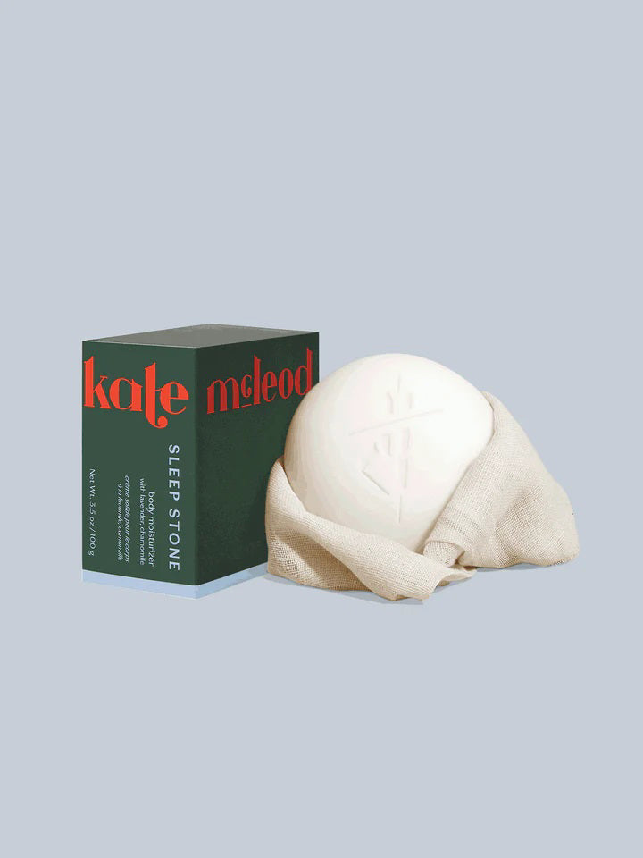 A smooth, round object wrapped in light beige lies beside a dark green box labeled "KATE MCLEOD" and "Sleep Stone," hinting at lavender essence. The moisturizing lotion bar refill and packaging sit elegantly against a light gray backdrop.