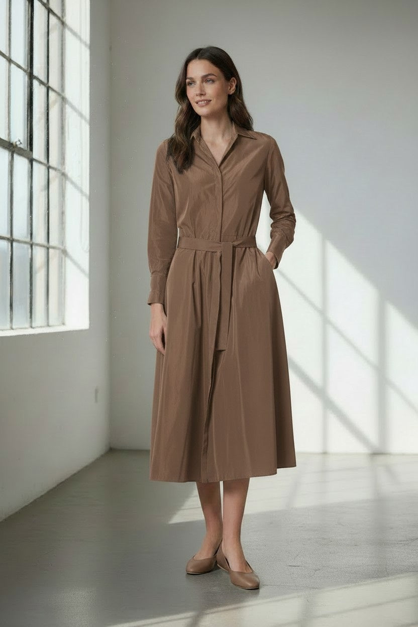 A woman stands in a sunlit room, her hands in her pockets and a slight smile on her face, wearing the VICARIO CINQUE ANNA Taffeta Shirtdress in Light Taupe.