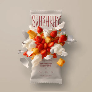 A STARS+HONEY - STRAWBERRY SHORTCAKE Collagen Protein Bar wrapper bursts open, releasing strawberries, cake pieces, and swirls of whipped cream against a neutral background—perfectly capturing the delicious essence of this wholesome treat. Enjoy all the flavor you love in strawberry shortcake, now in a nourishing STARS+HONEY Collagen Protein Bar.