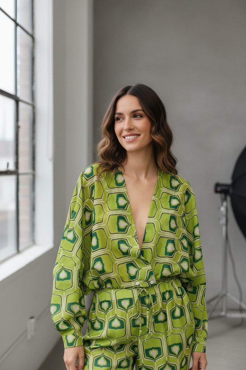 A woman with wavy brown hair wears the FLOOR - PRINTED BLOUSE by FLOOR, standing by a large window in a bright, modern room with gray walls and photography equipment in the background.