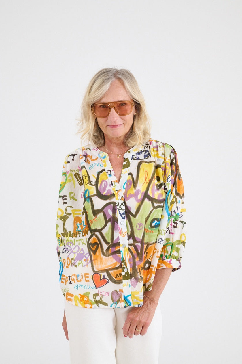 A blonde woman in sunglasses wears the STORIATIPIC - COCO LOVE BLOUSE IN WHITE, featuring colorful graffiti-style designs, standing with her back to the camera against a plain white background for a look of casual elegance and comfort.